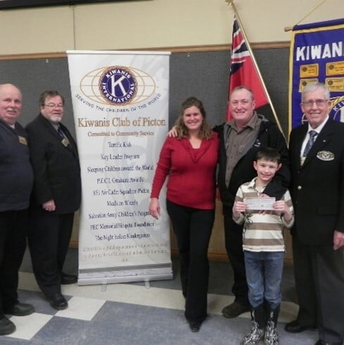 Two owners of Merland Park along with a child present their donation to three members of the Kiwanis in front a Kiwanis banner and an Ontario flag.