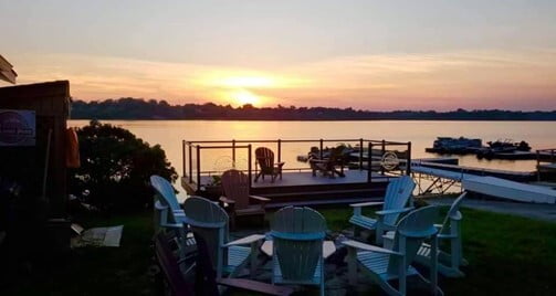 Chairs sit around a fire pit on a dock, while the sun is setting.