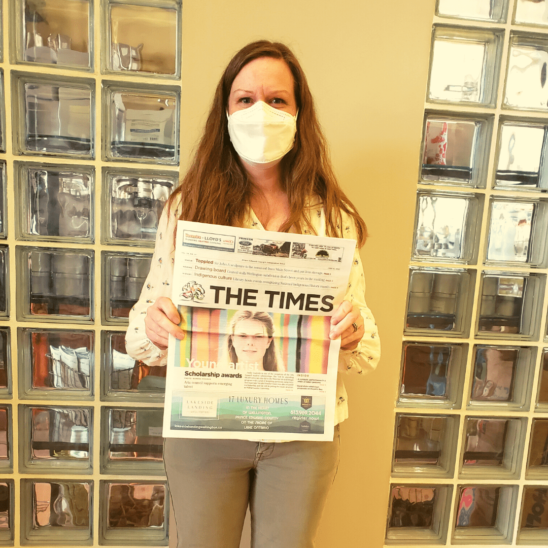 Executive Director of PEC Arts Council proudly holding up the front page of a newspaper that showcases an article about the PEC Arts Council grants for youth