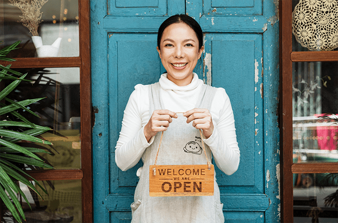 Business owner standing infront of the door to her business holding a sign stating "open" and smiling